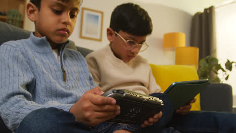 Two-Young-Boys-Sitting-On-Sofa-At-Home-Playing-Games-Or-Streaming-Onto-Digital-Tablet-And-Handheld-Gaming-Device